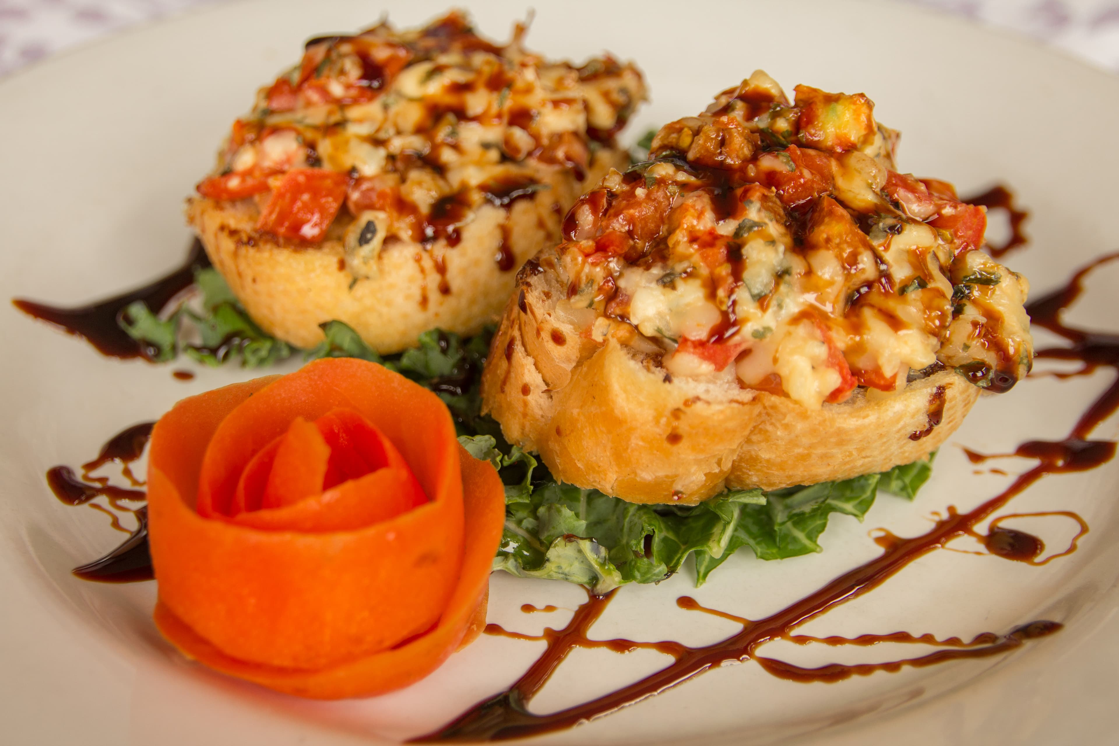 Two stuffed bread rolls topped with vegetables and drizzled with sauce, accompanied by a carrot rose on a bed of greens.