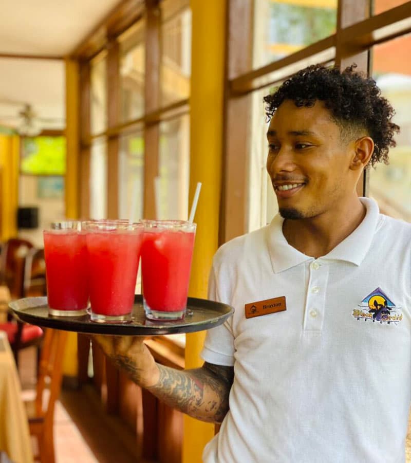 A smiling server holds a tray of four pink drinks in a sunny restaurant setting.