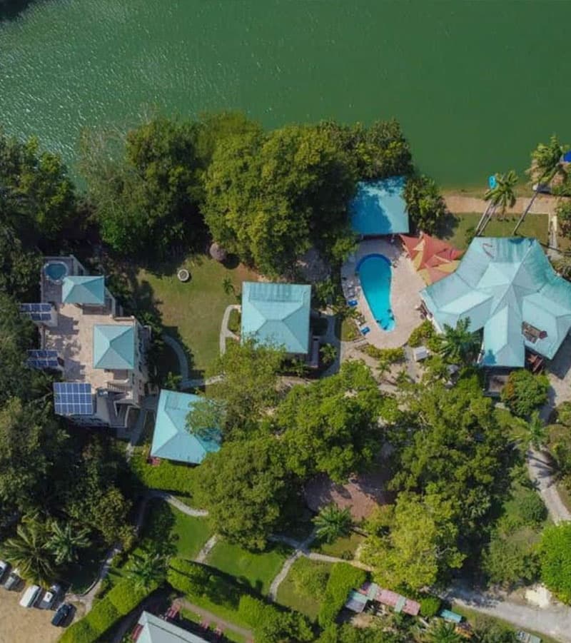 Aerial view of a tropical property featuring several buildings, a swimming pool, and lush greenery adjacent to a body of water.