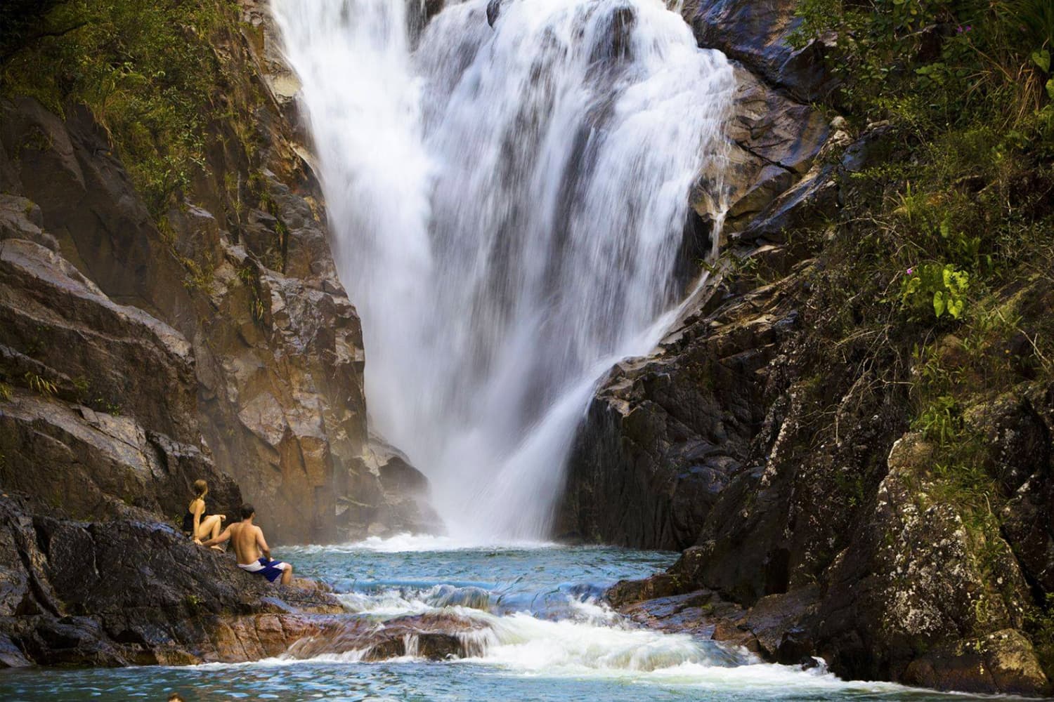 A person sits on rocks by a waterfall cascading into a blue pool.