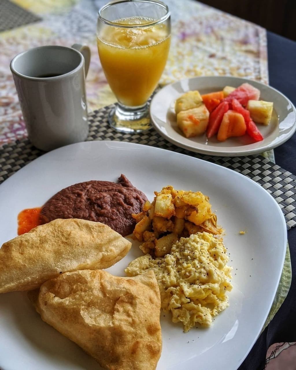 A plate of breakfast with two fried pastries, scrambled eggs, potatoes, and refried beans, accompanied by a fruit bowl and beverages.