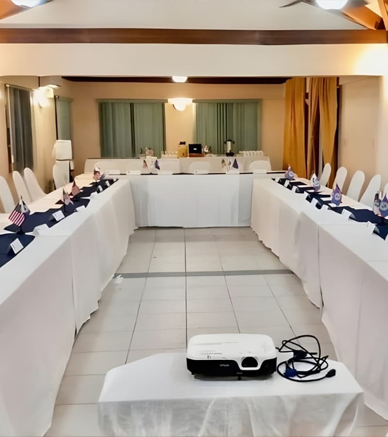 A conference room setup with white tables, chairs, and flags, featuring a projector at the front.