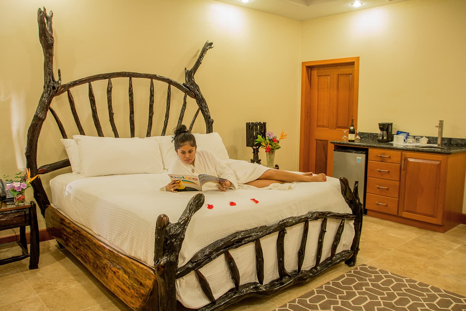 A woman in a robe reading a book on a rustic bed in a warmly lit hotel room.