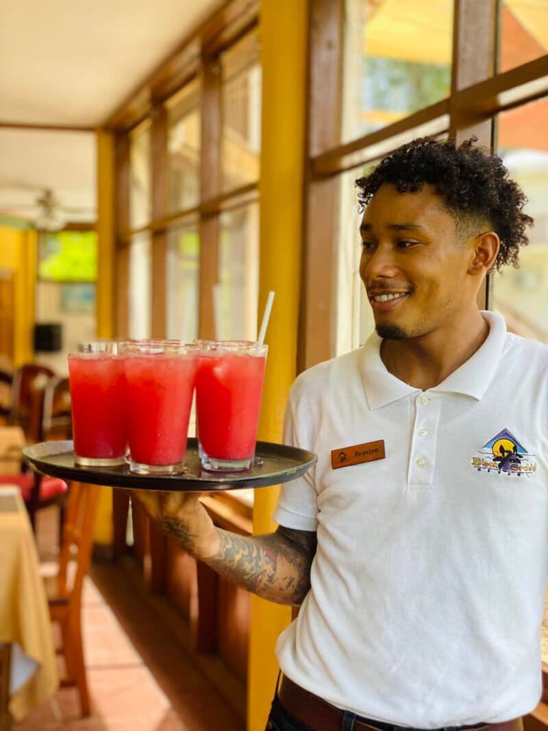 A smiling server carries a tray of pink drinks in a sunny restaurant.